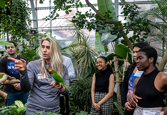 Link and photo of Stephanie Witte giving a tour to a group of visiting college students.  A tropical bird is in one of her hands and she is pointing out an interesting plant.